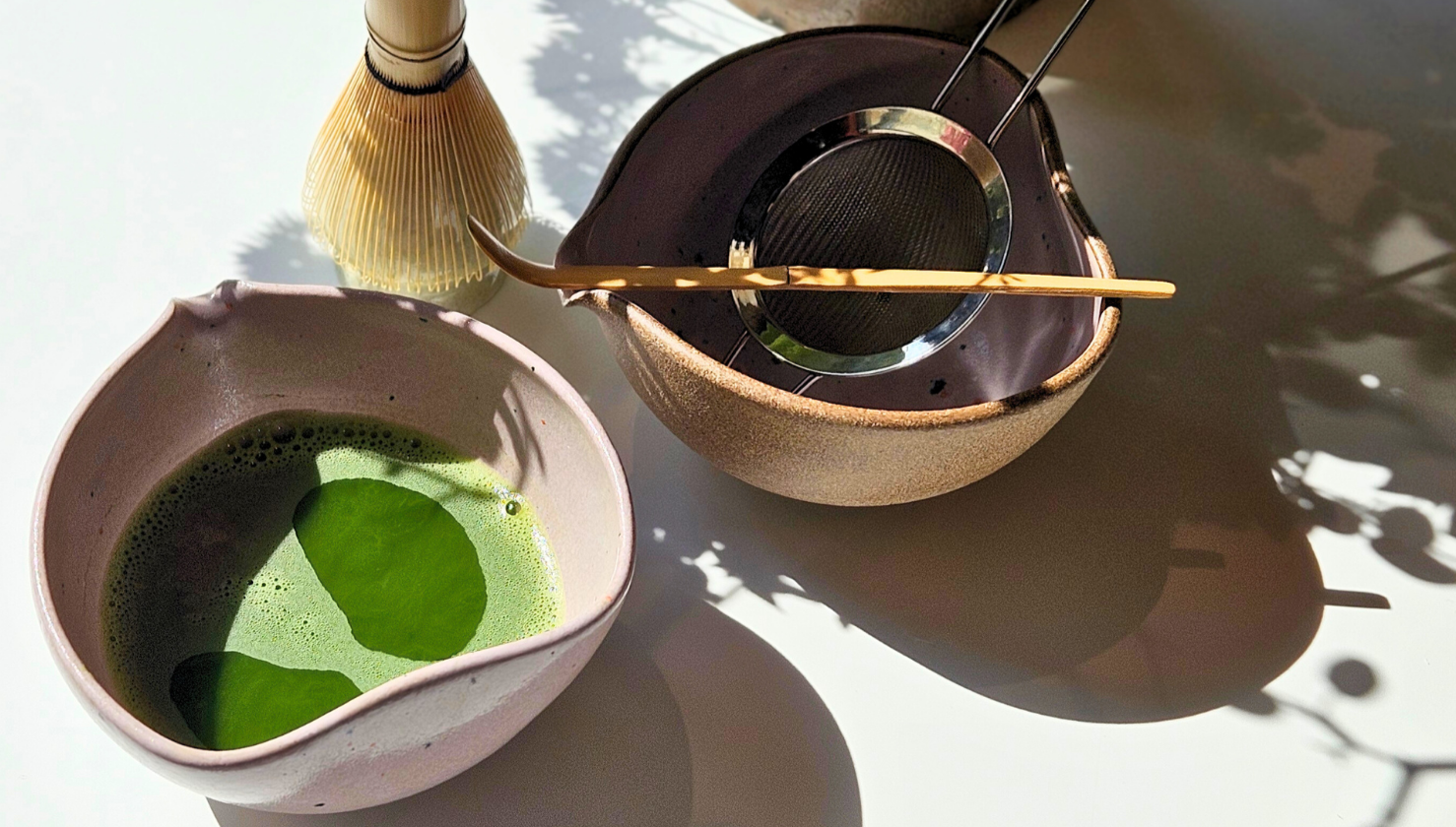 Matcha green tea preparation with a bowl of matcha, whisk, and other tools on a light surface.